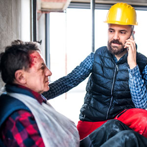 A construction worker with a head wound is being assisted by a coworker on a job site.